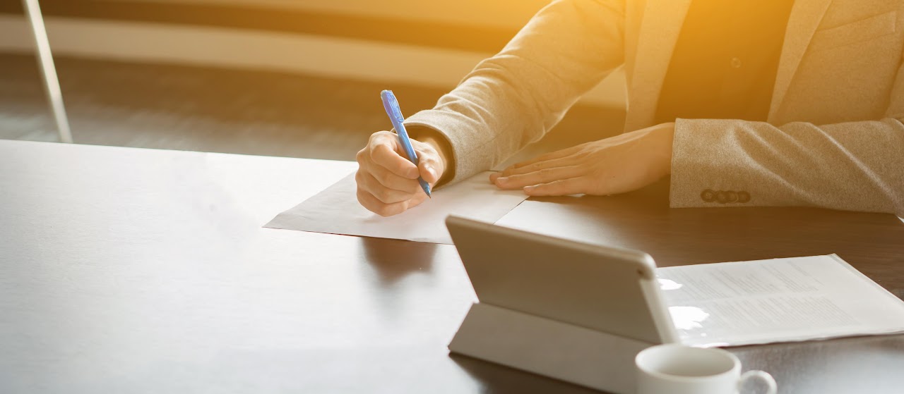Person writing notes on paper at a desk with a tablet nearby.
