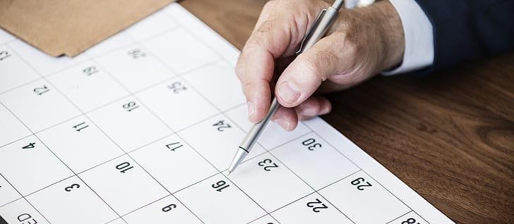 Hand holding a pen pointing to a specific date on a paper calendar.